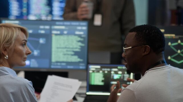 Zoom out shot of diverse team of network engineers communicating while working in control center with programming codes and software on monitors