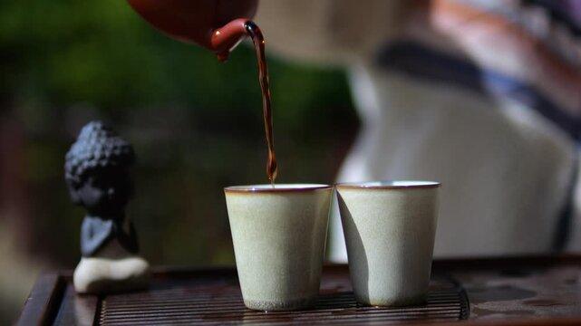 Close up of a person pouring hot black tea from a traditional brown clay teapot into two small ceramic cups during a relaxing and mindful outdoor asian gongfu cha ceremony in a sunlit garden