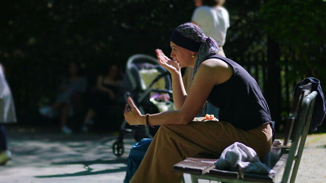 Woman undergoing chemotherapy on park bench during video call, smiling while listening and reacting with expressive gesture in warm sunlight