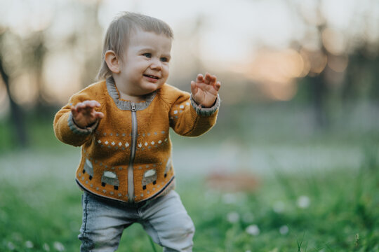A smiling toddler in a mustard sweater takes tentative steps on green grass. Warm family moment suggesting kids and mom enjoying the outdoors.
