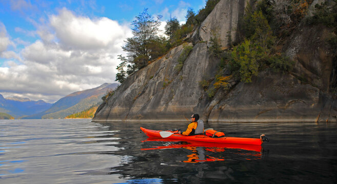 View of a man in a red kayak paddling on the calm waters of Nahuel Huapi Lake by steep rocky cliffs San Carlos de Bariloche, Rio Negro, Argentina.