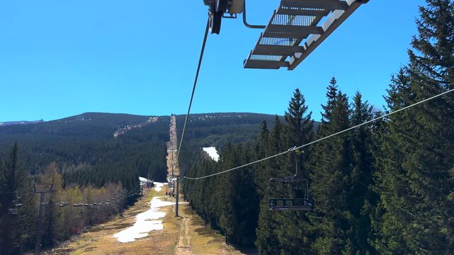 First person perspective riding a chairlift up a scenic mountain, showing melting snow on the ski slope below and a dense pine forest on a beautiful sunny day in early spring