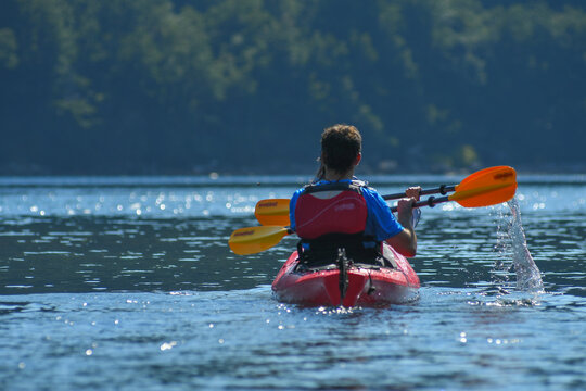 View of a person paddling a red kayak with orange oars on the calm blue waters of Nahuel Huapi Lake with lush green forests in the background San Carlos de Bariloche, Rio Negro, Argentina.
