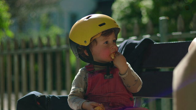 Toddler in yellow helmet seated on park bench, nibbling tangerine slices in soft sunlight, candid moment of quiet focus and sensory discovery during outdoor play