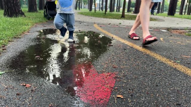 Baby boy walking through puddles in a park wearing rubber boots together with his father. 4k, slow motion.