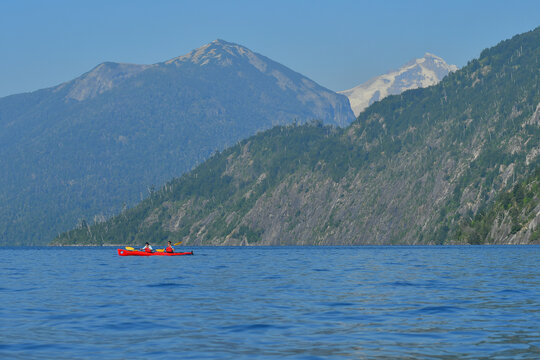 View of two people in a red kayak on the calm waters of Nahuel Huapi Lake with steep forested mountains and a clear blue sky in the background San Carlos de Bariloche, Rio Negro, Argentina.