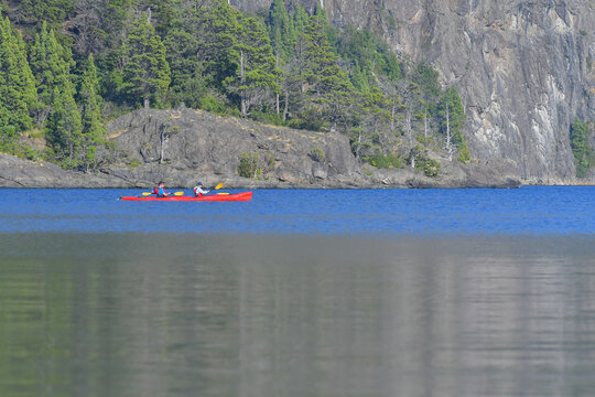 View of two people paddling a red kayak on the calm blue waters of Nahuel Huapi Lake with rocky shores and green trees SAN CARLOS DE BARILOCHE, Rio Negro, Argentina.