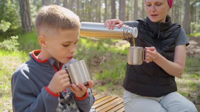 Mother pouring hot drink from thermos flask for son in woods. Parent fills metal mug with warm beverage on bench during family hike.