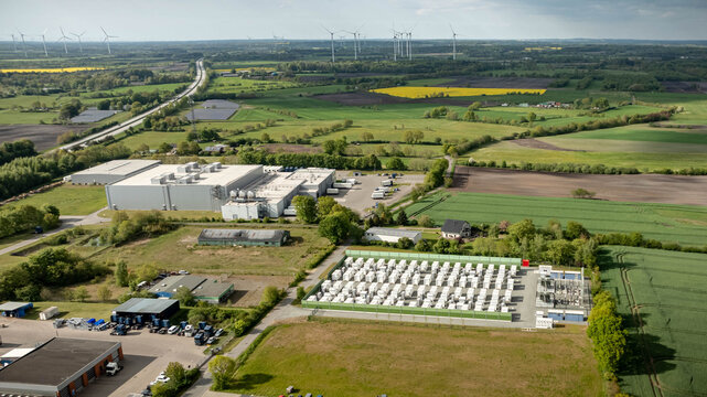 Aerial view of industrial energy storage facility by agricultural fields and renewable energy landscape with solar panels and wind turbines in the countryside. Battery energy storage system (BESS)