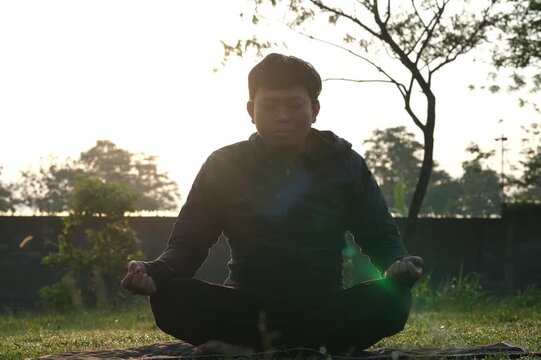 A man sits cross-legged doing meditation and yoga, breathing fresh air, relaxing his body in the park with the silhouette of the soft morning sunlight.