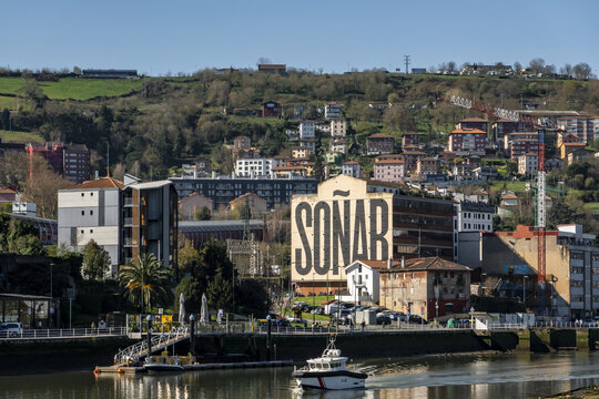 View of the Nervion river with a boat passing by the SONAR mural and hillside residential buildings under a clear sky in Greater Bilbao, Basque Country, Spain.