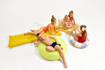 Obraz premium Children relaxing on colorful inflatable pool floats in white studio with large copy space. Concept of summer camp promo, kids swimwear ads, family travel offers and pool toy marketing.