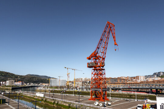 View of the iconic red Carola Crane standing on the waterfront of the Nervion River with city buildings and a clear blue sky Greater Bilbao, Basque Country, Spain.