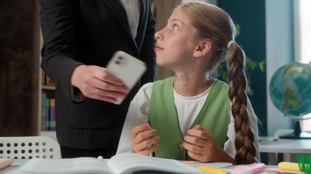 Caucasian little girl child playing mobile phone game at class lesson in school woman teacher female tutor taking away smartphone continue play online gaming gadget addict pupil kid confused writing