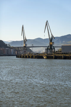 View of industrial cranes standing on the waterfront of the Nervion river with mountains and a bridge in the background in Greater Bilbao, Basque Country, Spain.