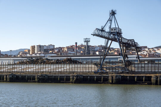 View of a large blue industrial crane and piles of scrap metal at the port with the city skyline in the background Greater Bilbao, Basque Country, Spain.