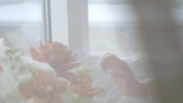 A close-up of a delicate bride in a white silk negligee, sitting on the windowsill and touching a bouquet of flowers with her hands. The video was shot through a transparent cloth. 