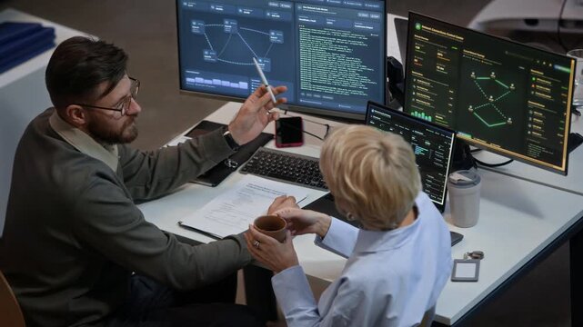 View from above of two Caucasian engineers discussing and analyzing network system while sitting at table with computers in cybersecurity office