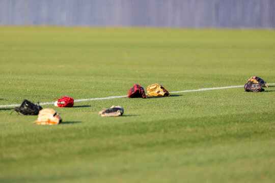 Multiple baseball gloves and baseballs rest on the grass beside a painted field line during pregame warmups at a high school baseball game in Olive Branch, Mississippi.