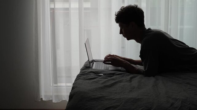 Digital nomad finishing online work on laptop and falling back onto bed in apartment with large window, remote job fatigue, deadline pressure and recovery after work
