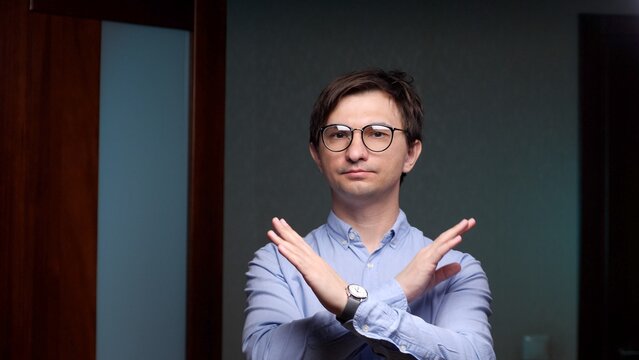 Young man in round glasses and blue shirt forms an x with crossed arms, conveying refusal, stop, boundary setting, disapproval and firm objection in a studio portrait