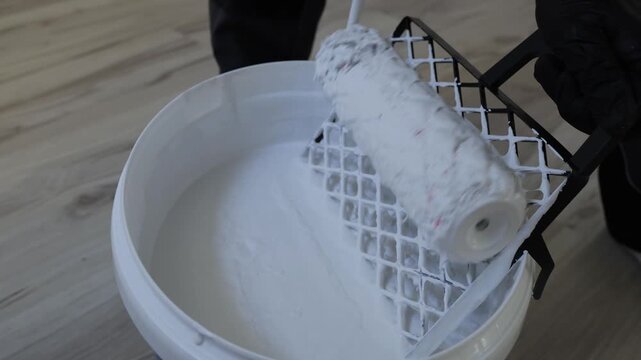 Close up of paint roller covered in white paint inside bucket during apartment renovation and interior wall painting process with professional decorating tools