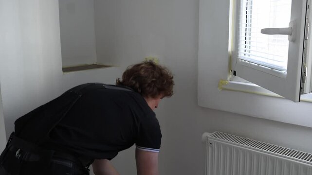 Young man painting apartment wall near bright attic window with roller while renovating modern home interior during residential repair and decorating work