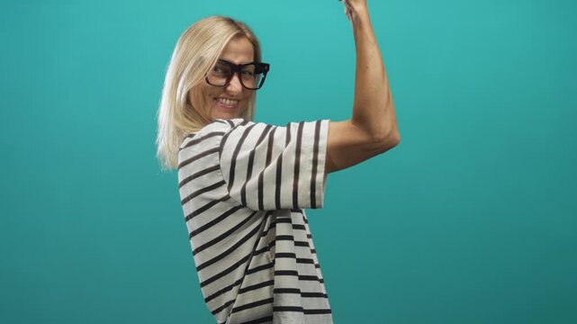 Woman flexes arm showing bicep and smiling while wearing striped shirt in studio teal backdrop; confidence empowerment.
