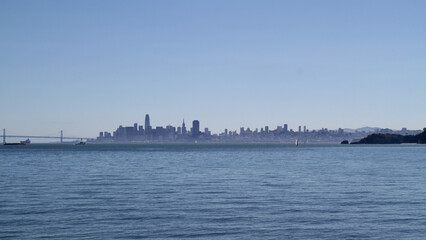Naklejka premium San Francisco city skyline silhouette seen from the bay at golden hour, with the Bay Bridge visible on the left and calm waters in the foreground, California, USA.
