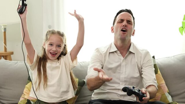 A father and daughter sitting on a sofa playing video games together, both holding controllers and focused on the screen.