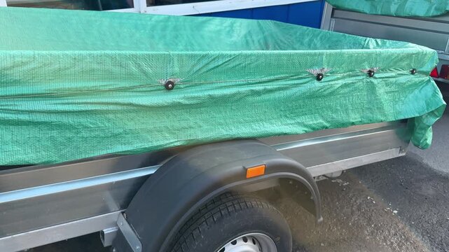 Small car trailer covered green tarp, parked roadside on asphalt near dealership, visible wheel, fender and hitch, light rust