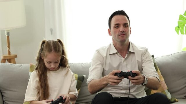 A father and daughter sitting on a couch playing video games together, both holding controllers and appearing engaged.