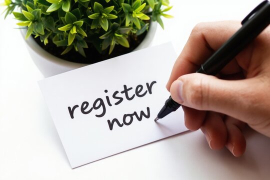 Hand writes register now text on paper card. Green plant grows in pot nearby. Call to action concept. Plan to sign up. Make decision now. Complete form online.
