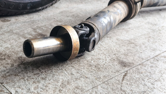 Close-up of a used, greasy metal automotive driveshaft with a universal joint and splined slip yoke resting on a concrete floor
