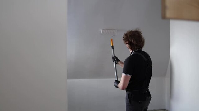 Young man carefully repainting attic apartment walls with roller during budget home renovation while standing alone in unfinished empty room with fresh white paint