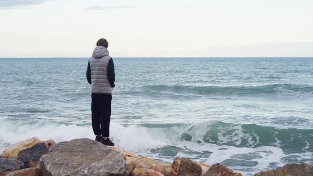 Lonely boy sitting on seaside rocks watching restless waves under overcast sky. Emotional solitude, introspection, and mental reflection concept by the ocean.