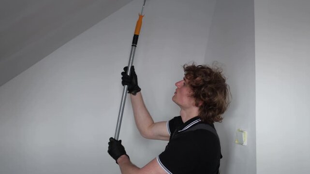 Young apprentice painter learning apartment renovation skills while repainting attic walls with roller during practical construction training in empty room