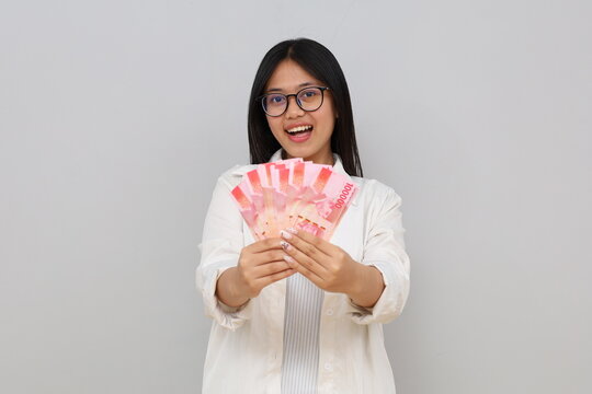 Portrait of a young Asian woman wearing casual clothes, holding Indonesian rupiah banknotes, showing money forward, winning the lottery, standing isolated on white background.
