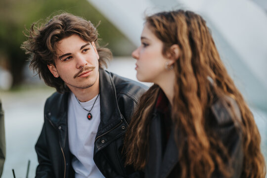 Two high school friends sit together outdoors in casual leather jackets and converse. A young man listens attentively while his classmate looks away in thought.