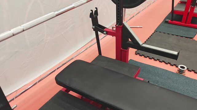 Weight bench setup in a gym with black padded seat and red metal frame, barbell rack positioned above, rubber flooring and exercise mats visible in the background