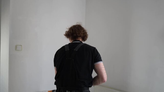 Young man repainting apartment wall from behind during home renovation project applying fresh white paint in empty room with clean minimalist interior atmosphere