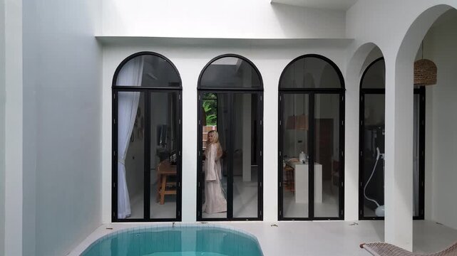 Blonde Woman Walking Inside Luxury Tropical Villa Behind Panoramic Windows
