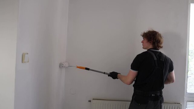 Young man repainting apartment walls with roller during home renovation project creating clean fresh interior before moving into new living space