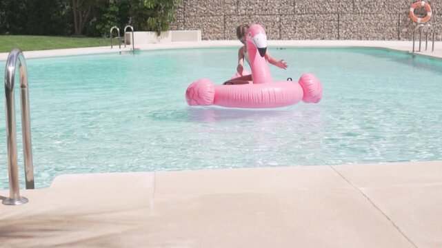 Little girl sitting on a pink inflatable flamingo in a bright outdoor swimming pool. sstkSummer lifestyle video showing sunny vacation, childhood water play, pool recreation and relaxed summer fun