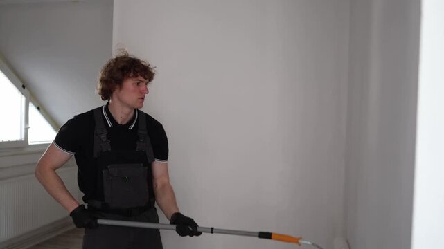 Young beginner painter carefully rolling fresh white paint on apartment wall during first home renovation experience in empty room with natural daylight atmosphere