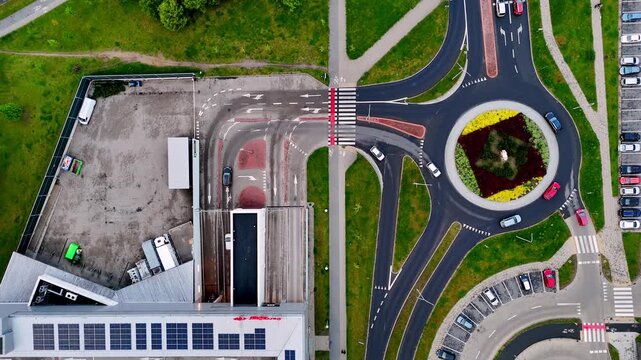 Drone film showing car traffic at a shopping mall, traffic roundabout, parking lot with vehicles, and building roof with solar panels, featuring road markings, pedestrian crossings, and green lawns fr