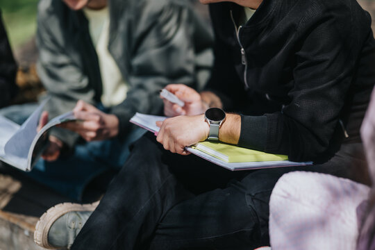 A group of school friends sit outdoors reading notebooks and taking notes. The students study together in a relaxed, collaborative setting.