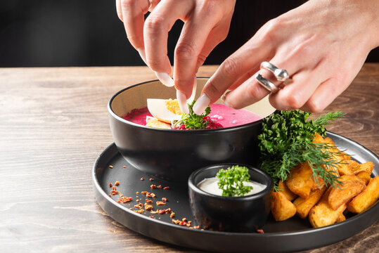 A person's hands garnish a bowl of pink beetroot soup served with boiled egg, sour cream, and roasted potatoes on a wooden table.