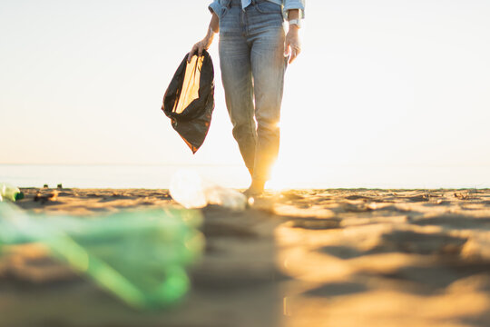 Person walking on a sandy beach cleaning up plastic bottles and trash, collecting garbage in a black bag, promoting environmental protection and sustainability at sunset