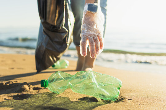 Volunteer cleaning beach, wearing plastic glove and holding trash bag, picking up plastic bottle from sand, protecting ocean environment from pollution